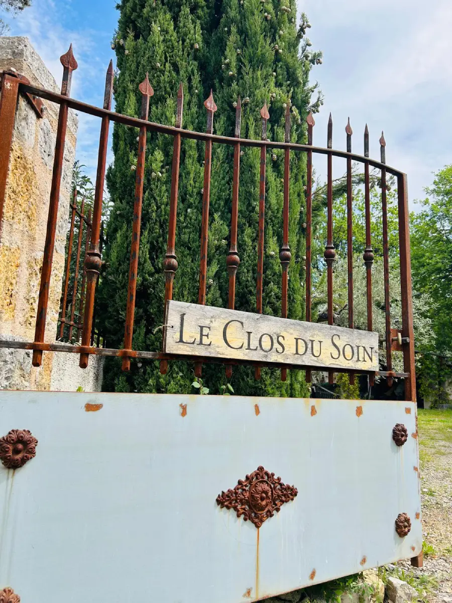 Plaque en bois « Le Clos du Soin » sur le portail en fer forgé, bordé de cyprès